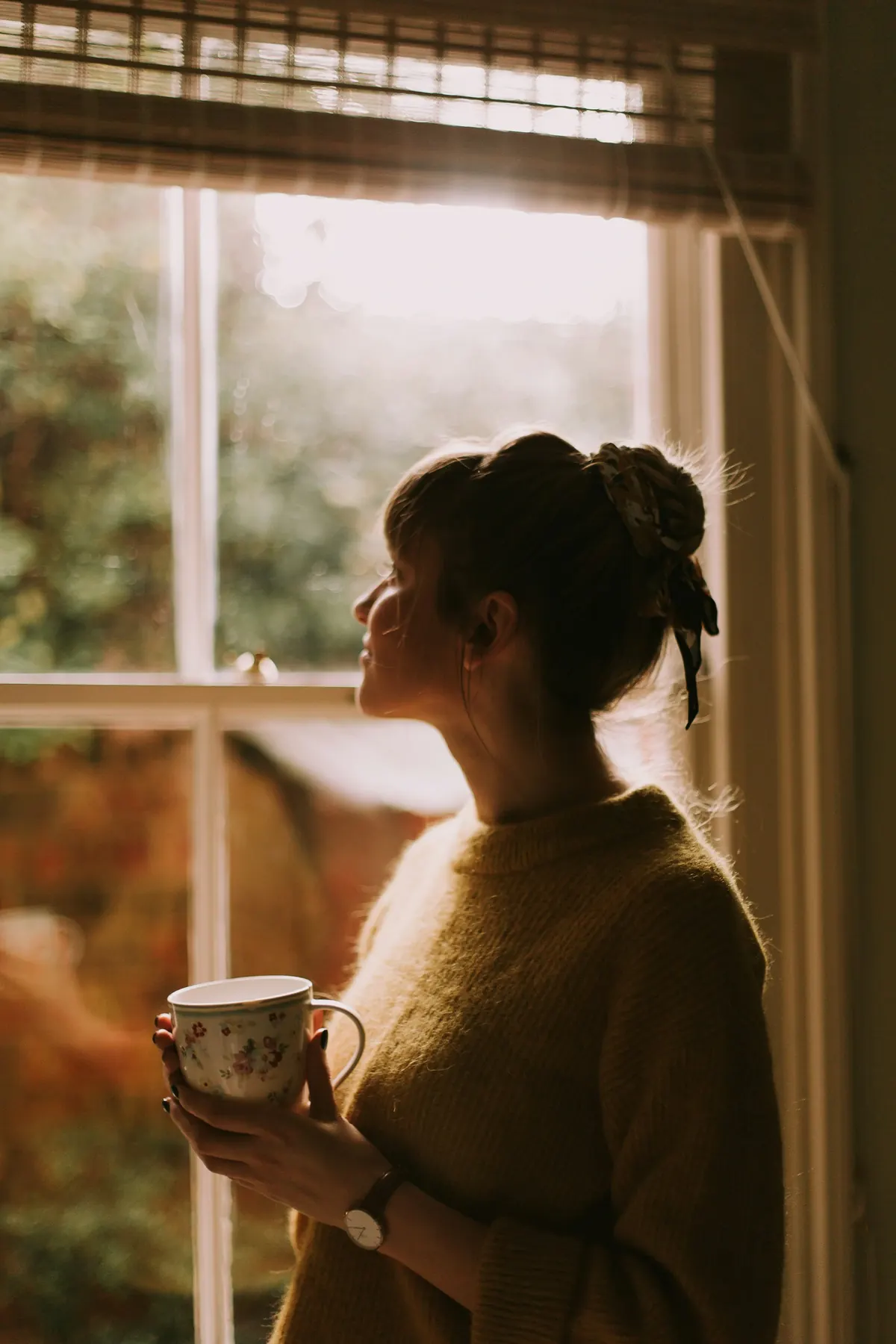 A woman holds a cup of tea by a sunny window in the afternoon.
