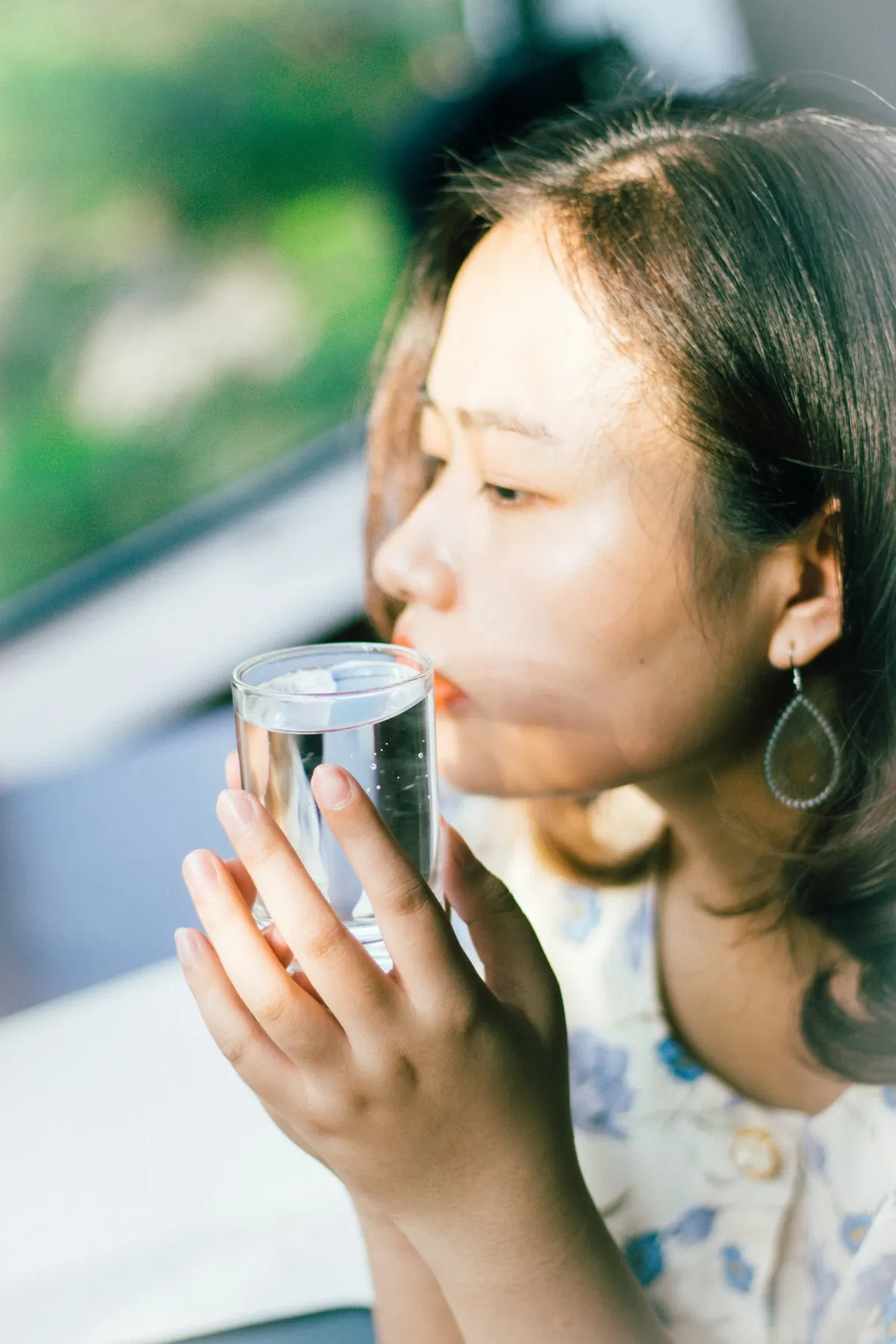 A woman sips a glass of water by a window.